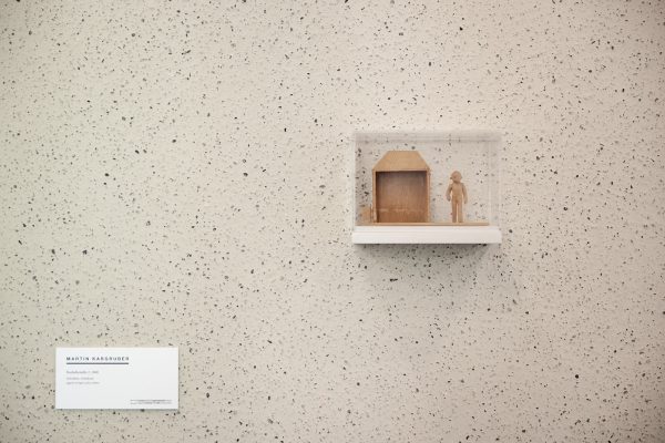 Objekt von Martin Kargruber, „Bushaltestelle 2“ (2000): kleines Holzmodell einer Haltestellenhütte mit stehender Figur in transparenter Vitrine auf weißem Regal vor gesprenkelter, heller Wand; links unten ein Werkschild.