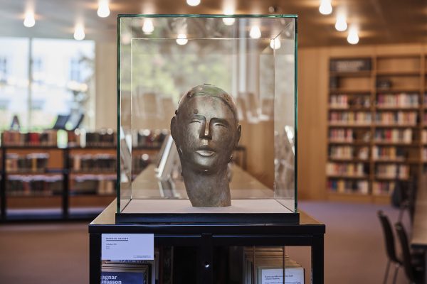 Skulptur von Markus Gasser, „Gedanken“ (2022): stilisierte Kopfplastik mit metallischem/bronzenem Eindruck in gläserner Vitrine auf schwarzem Sockel; im Hintergrund unscharfe Bücherregale und warmes Licht einer Bibliothek.